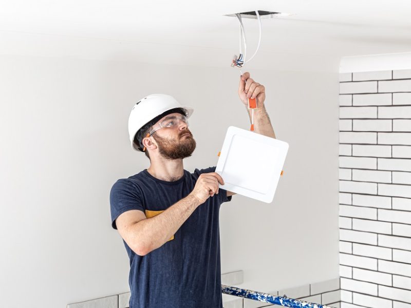 Electrician Builder with beard worker in a white helmet at work, installation of lamps at height. Professional in overalls with a drill on the background of the repair site.